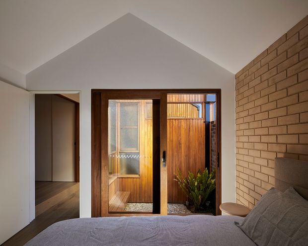 A warm, modern bedroom viewed toward a glazed timber-framed opening onto a small planted courtyard. The room has a pitched white ceiling, exposed tan brick feature wall, dark timber flooring, and a partially visible upholstered bed. A timber-lined outdoor area and a secondary interior space are visible beyond the opening.
