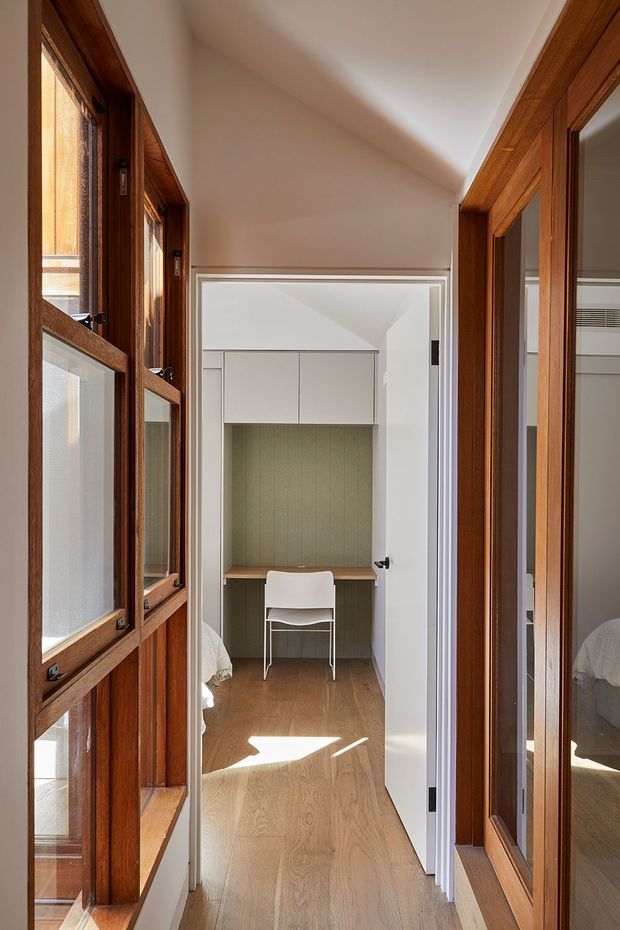 A narrow, sunlit interior passage leads to a compact built-in study nook. Warm timber-framed windows and glazed wardrobe or door panels line both sides, while the nook at the end has a wood desk, pale built-in overhead cabinetry, muted green vertical wall panelling, and a simple white chair. Light timber flooring and white walls create a calm, warm space.