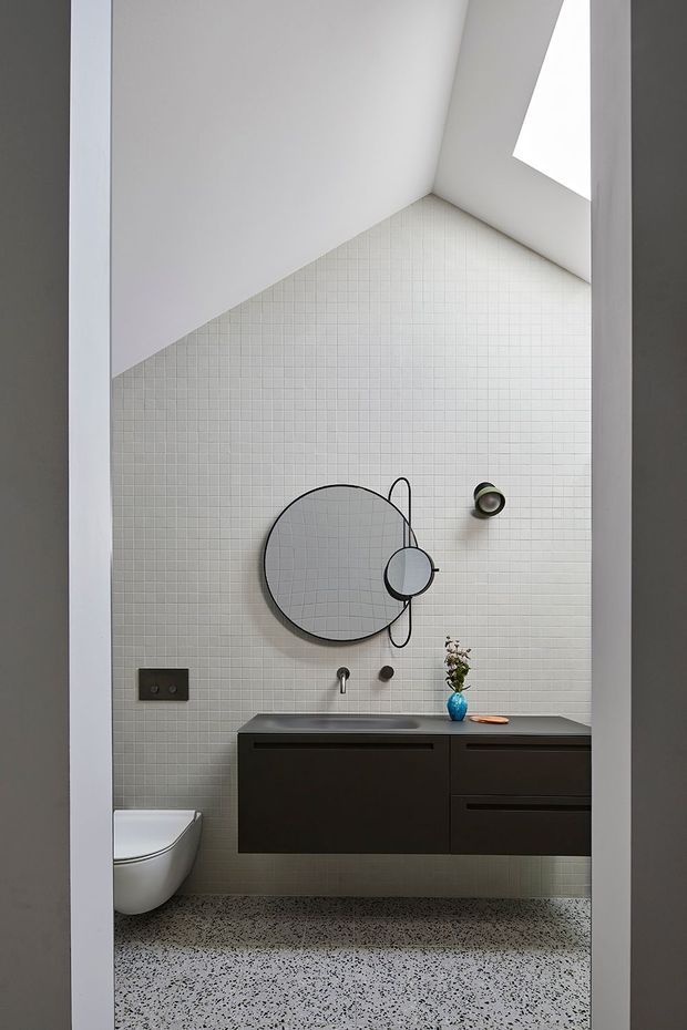 A compact contemporary bathroom viewed through a doorway, featuring full-height white micro-mosaic tiled walls, a speckled terrazzo floor, a dark floating vanity with integrated basin, wall-mounted tapware, a white wall-hung toilet, round layered mirrors, and a high angled skylight or window. The restrained monochrome palette and sculptural mirror arrangement create a clean, refined interior.