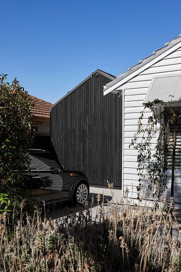 Exterior view of a compact residential property in bright daylight, showing a white weatherboard house with a louvred window shutter, an adjacent dark vertical-boarded gabled structure, and a narrow gravel-paved driveway. A black car and established planting occupy the foreground.