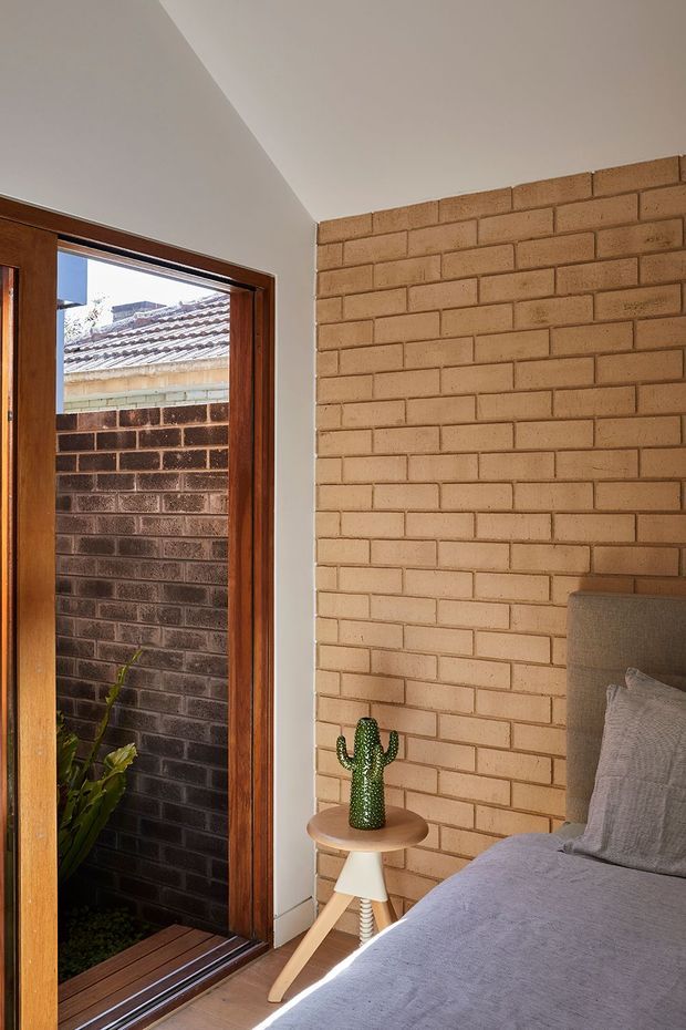 A compact bedroom corner with an exposed warm tan brick feature wall, a timber-framed glazed door opening to a small planted courtyard, and a partially visible upholstered bed with neutral bedding. A small round side table holds a cactus, while sunlight enters through the doorway and creates a warm, tactile atmosphere.