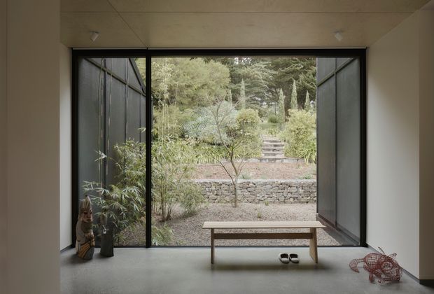 A minimalist interior opening onto a landscaped courtyard garden. Full-height black-framed glazed doors are opened on both sides, creating a broad connection to the exterior. The room has smooth pale walls and ceiling, a polished grey concrete floor, a simple timber bench, and sparse sculptural decor. Outside are gravel, planted trees and shrubs, stone retaining walls, and garden steps.