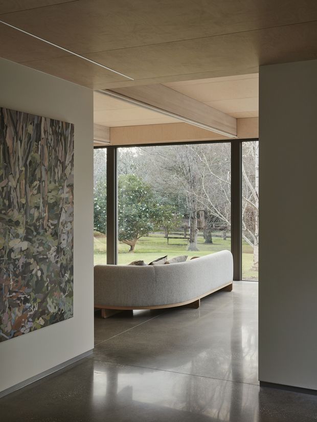 A refined contemporary living area viewed from an adjacent interior space, with a curved upholstered sofa positioned beside expansive black-framed glazing overlooking a landscaped garden. The room features a polished concrete floor, warm timber ceiling and soffit panels, slim linear ceiling detailing, pale neutral walls, and a large abstract artwork.