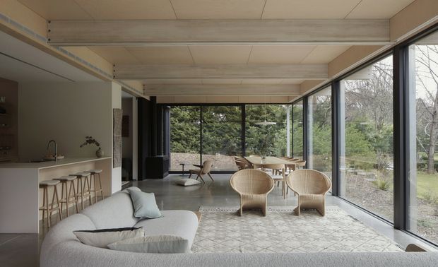 A refined open-plan living space with a compact kitchen at left, a pale upholstered sofa in the foreground, and a dining area surrounded by floor-to-ceiling glazing. Natural timber-toned ceiling panels and beams, dark window frames, woven lounge chairs, and a neutral patterned rug create a calm connection to the landscaped garden outside.
