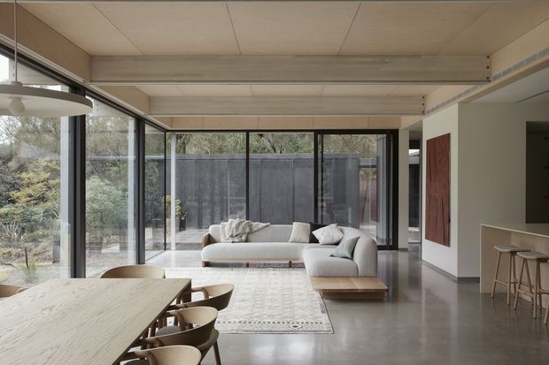 A refined open-plan living and dining space with floor-to-ceiling black-framed glazing opening to a landscaped courtyard. The room features a large timber dining table with curved chairs, a light sectional sofa, patterned area rug, low timber coffee table, and a compact kitchen or bar area at right. Pale timber ceiling panels and exposed structural beams contrast with polished grey concrete flooring and neutral plaster walls. The overall aesthetic is calm, contemporary, and connected to the outdoors.