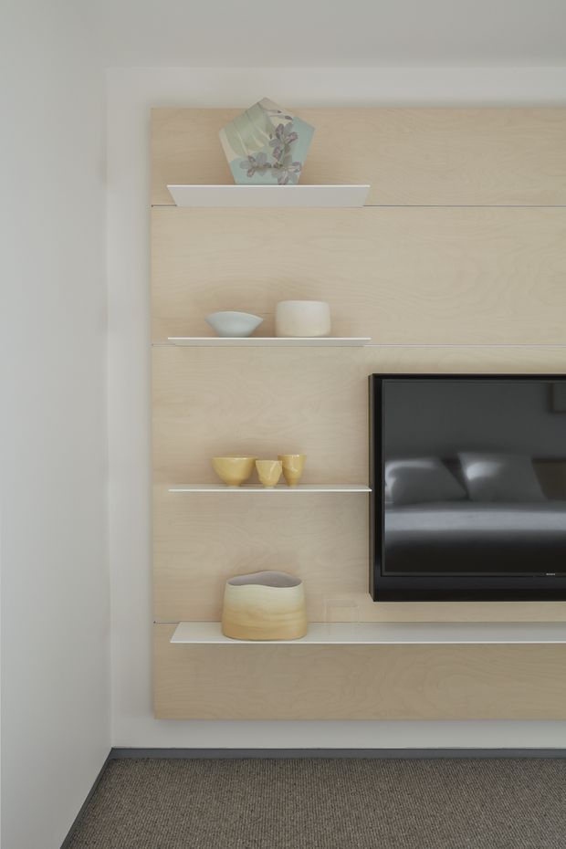 A minimalist living room media wall featuring a large pale timber veneer panel with slim white floating shelves and a long white display shelf. Decorative ceramics and a vase are arranged on the shelves, with a wall-mounted black television on the right. The room has white painted walls, grey carpet, and clean, restrained detailing.