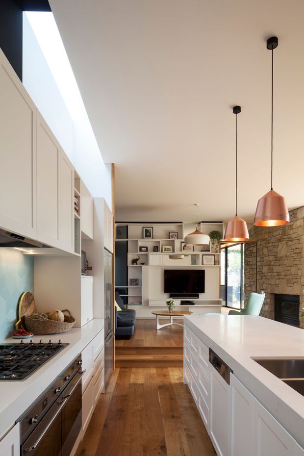 A narrow, open-plan kitchen looking toward an adjoining living room. The kitchen has pale shaker-style cabinetry, a white island with an integrated sink, a gas cooktop and stainless-steel oven, warm timber flooring, and a pale blue geometric-tile splashback. Three copper pendant lights hang over the island. The living area beyond includes built-in white shelving around a television, a dark sofa, and a substantial exposed-stone fireplace wall. A skylight or clerestory opening brings in strong natural light.