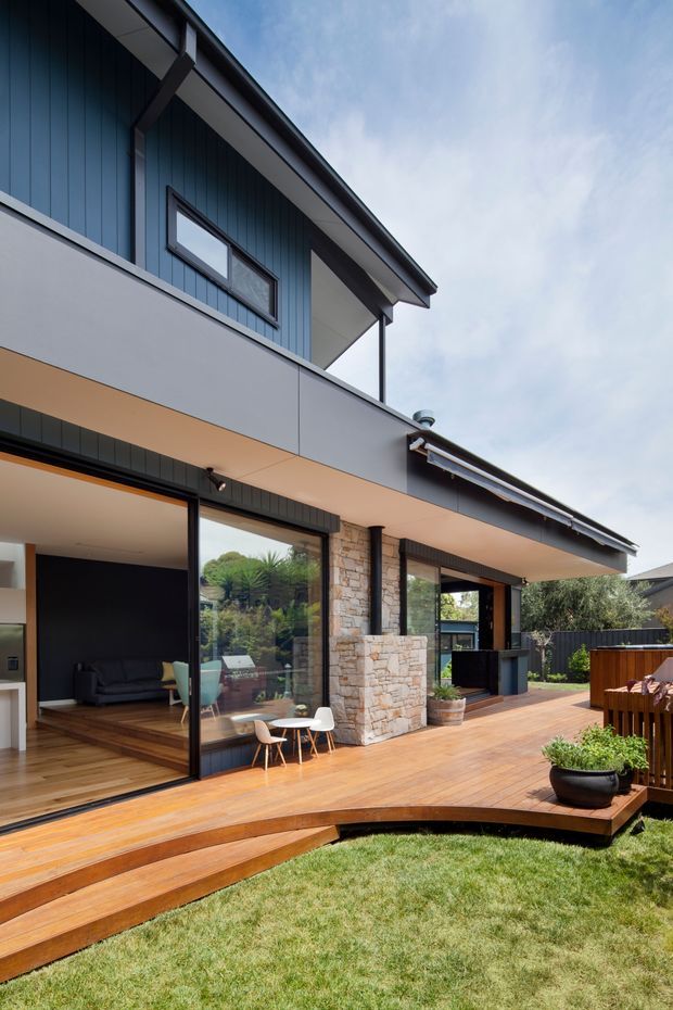 Exterior view of a contemporary multi-level home opening onto a broad timber deck and landscaped lawn. The facade combines dark blue vertical cladding, pale smooth panels, large black-framed sliding glass doors, and a prominent rough stone-clad chimney or fireplace element. Curved timber steps connect the deck to the grass, while an outdoor kitchen or barbecue area is visible at the right. Through the glazing, a living room is partially visible.