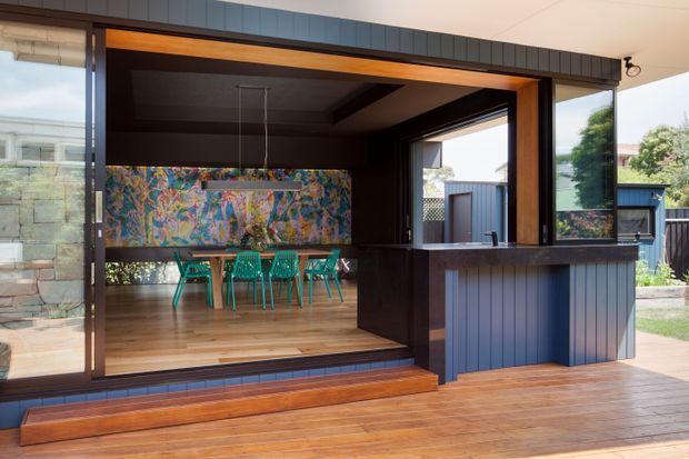 A modern indoor-outdoor entertaining area viewed from a timber deck. Large glazed folding or sliding doors open into a dining room with a colourful abstract artwork, turquoise dining chairs, and a timber table. The extension features dark vertical cladding, deep timber-framed openings, a dark ceiling, and a compact built-in bar or serving counter facing the deck. The space has strong indoor-outdoor connection and contemporary detailing.