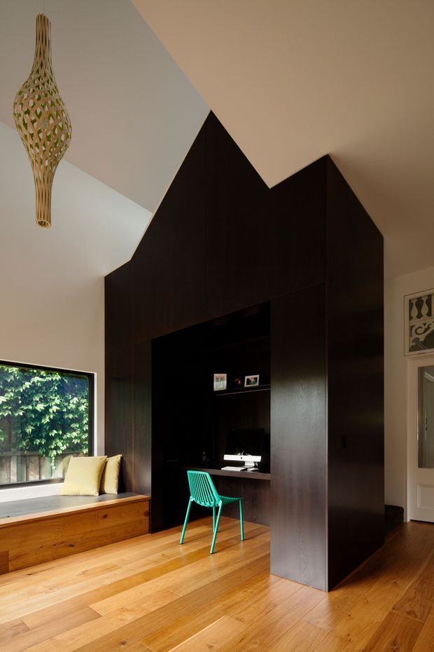 A compact, architecturally designed home office nook integrated into a tall, dark timber built-in enclosure with a steep house-like silhouette. The office contains a desk, computer and bright turquoise chair, while a timber window seat with cushions sits beside a large window. Warm timber flooring, white walls and ceiling, and a sculptural woven pendant create a refined contemporary interior.