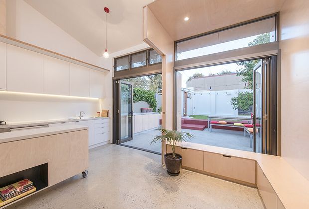 Bright contemporary kitchen opening onto a private courtyard through large glazed doors. The room features streamlined white full-height cabinetry, a pale timber kitchen island with integrated dark fireplace-like recess, light concrete flooring, pale timber window-seat joinery, exposed black-framed glazing, and minimal pendant and recessed lighting. Outdoor seating and planting are visible beyond the open doors.