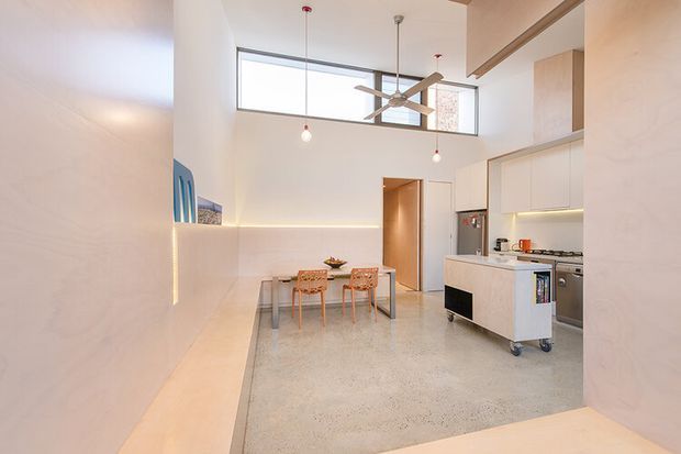 Bright open-plan kitchen and dining area with a polished concrete floor, white minimalist cabinetry, a compact island, built-in bench seating, and a small dining table. High clerestory windows and a ceiling fan create an airy, contemporary interior with warm concealed lighting and subtle timber accents.