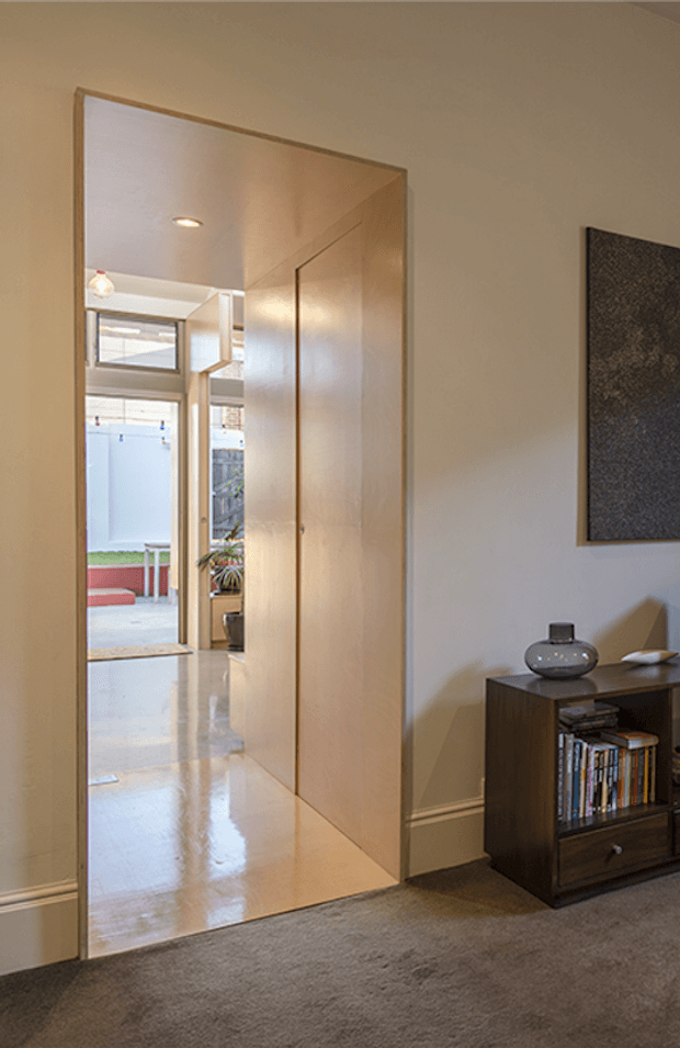 Interior view of a refined residential entryway or hallway, looking through a tall timber-trimmed opening toward a brighter courtyard or outdoor area. The space features warm neutral painted walls, glossy pale flooring extending through the passage, carpet in the foreground, full-height timber built-in cabinetry, and a dark timber low storage unit with books and decorative objects. A large framed dark artwork hangs on the wall, and daylight enters from the glazed opening beyond.