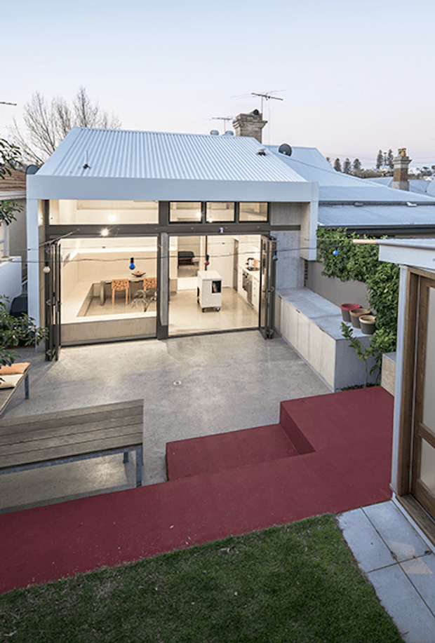 View from an upper-level outdoor area across a compact courtyard toward a modern glazed extension. The extension has a pale corrugated metal pitched roof, expansive dark-framed glazing and sliding doors, and a bright open-plan interior with a dining table and kitchen cabinetry visible inside. Concrete paving, raised planters, timber steps, and a burgundy-painted outdoor walkway frame the courtyard, with adjoining residential rooflines and chimneys in the background.