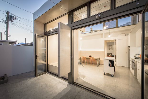 View from an outdoor courtyard or patio into a bright contemporary open-plan interior through large black-framed glazed sliding doors. The space contains a compact dining area, a kitchen with white cabinetry, a mobile storage unit, polished concrete floors, white walls, exposed ceiling services, and pendant lighting. The exterior includes a concrete-paved surface, privacy wall, and a covered overhang.