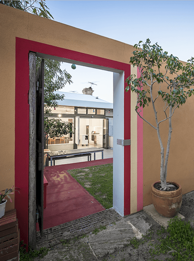 A view through a bold red-framed opening in a warm ochre rendered wall into a small landscaped courtyard and a contemporary residential interior. The foreground includes a dark timber gate, concrete paving, a potted tree, and a red pathway leading across grass toward the house. The house beyond has light rendered walls, extensive glazing, and a low-pitched metal roof.