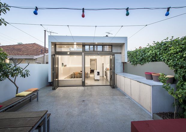 A contemporary rear courtyard or terrace viewed from outdoors, featuring a polished concrete-paved entertaining area, large dark-framed glazed doors, and a compact bright kitchen or living extension beyond. Low rendered boundary walls, built-in concrete planters, lush screening greenery, outdoor bench seating, and overhead festoon lighting create a private urban outdoor space. A neighbouring tiled roof is visible beyond the boundary.