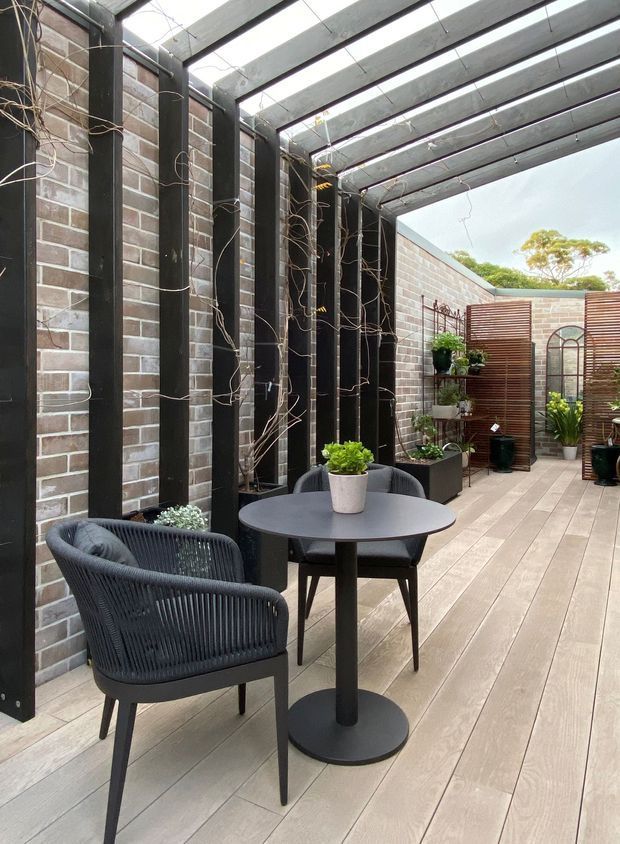 A narrow enclosed courtyard or terrace with pale brick walls, dark timber or metal vertical posts, and a translucent slatted pergola overhead. The space has light timber decking, black woven outdoor chairs, a round black café table, potted plants, climbing vines, and timber screening panels with decorative arch details. The overall design is a refined, contemporary outdoor entertaining space with strong linear structure and a muted neutral palette.