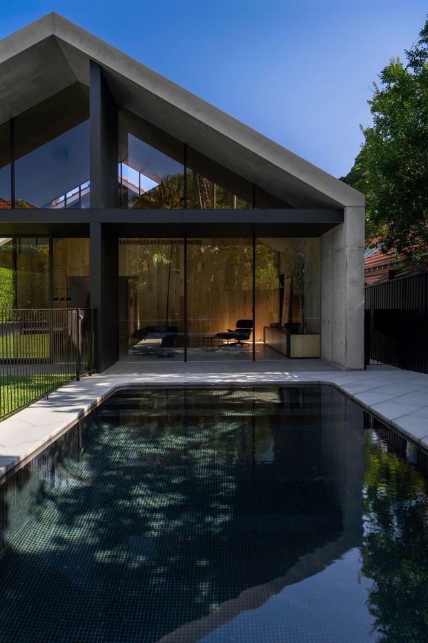 A modern concrete-and-glass house photographed from the pool area. The strong geometric facade has a dramatic sloping roof plane, dark structural framing, extensive floor-to-ceiling glazing, and warm timber interiors visible through the glass. A tiled rectangular swimming pool occupies the foreground, bordered by pale paving, with established trees and neighbouring residential elements around the site.