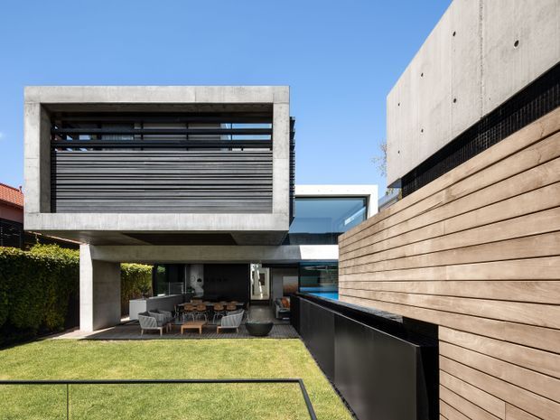 Contemporary multi-level residence viewed from a landscaped backyard, featuring a strong cantilevered concrete upper volume with horizontal louvres, extensive glazing, dark metal detailing, and a warm horizontal timber-clad boundary wall. The ground level opens to an outdoor dining and living area beside a lawn, with a glimpse of a pool or blue water feature.