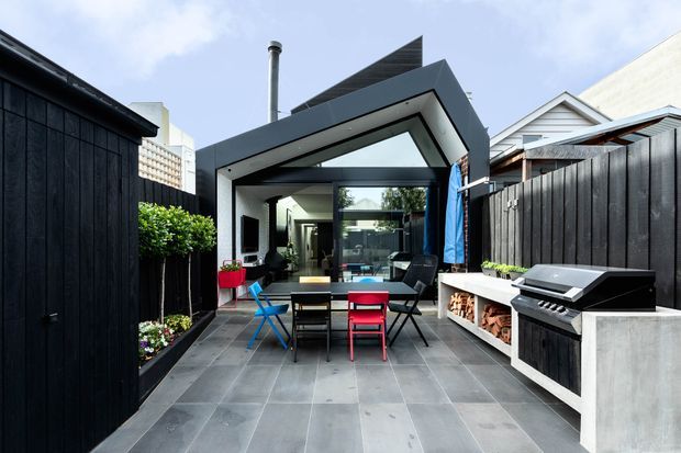 A contemporary courtyard entertaining area viewed from outdoors, featuring dark vertical timber fencing, large-format grey tiled paving, a covered glazed extension with a dramatic angular roofline, outdoor dining furniture, and a built-in barbecue bench with integrated firewood storage. The space connects visually to an interior living area through full-height glazing.