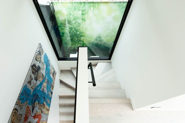 Bright modern stairwell with pale timber treads, white walls, a simple white balustrade and black handrail. A large overhead skylight brings in daylight and features a translucent green botanical graphic, while a colourful artwork leans against the stair wall.