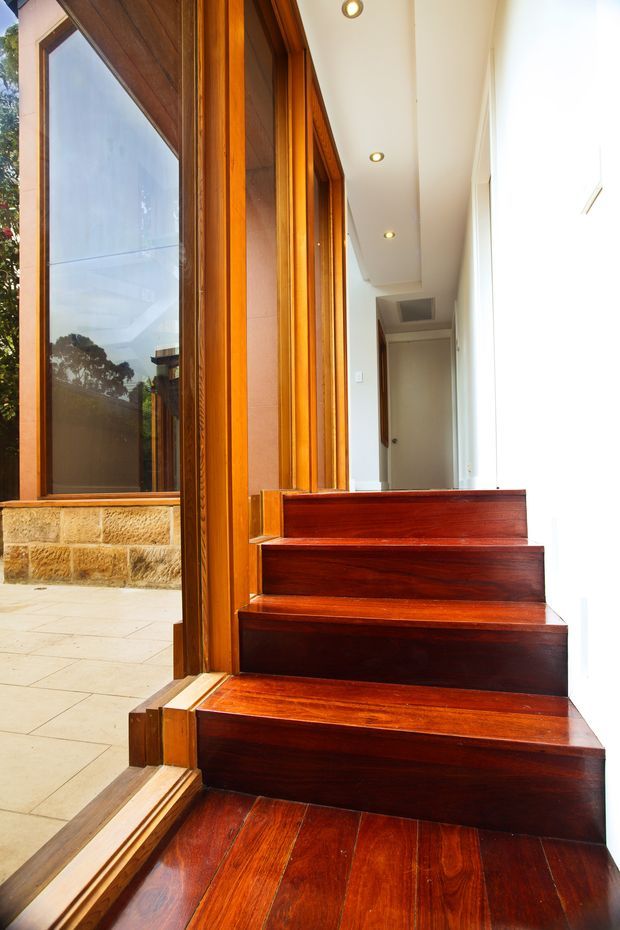 A bright interior stairway with richly finished dark timber treads and risers leading up alongside a large timber-framed glazed opening. The stair edge and surrounding joinery use polished warm-toned wood, while the adjacent corridor has white walls and recessed ceiling lights. Through the glazing, a stone-clad exterior wall and trees are visible.