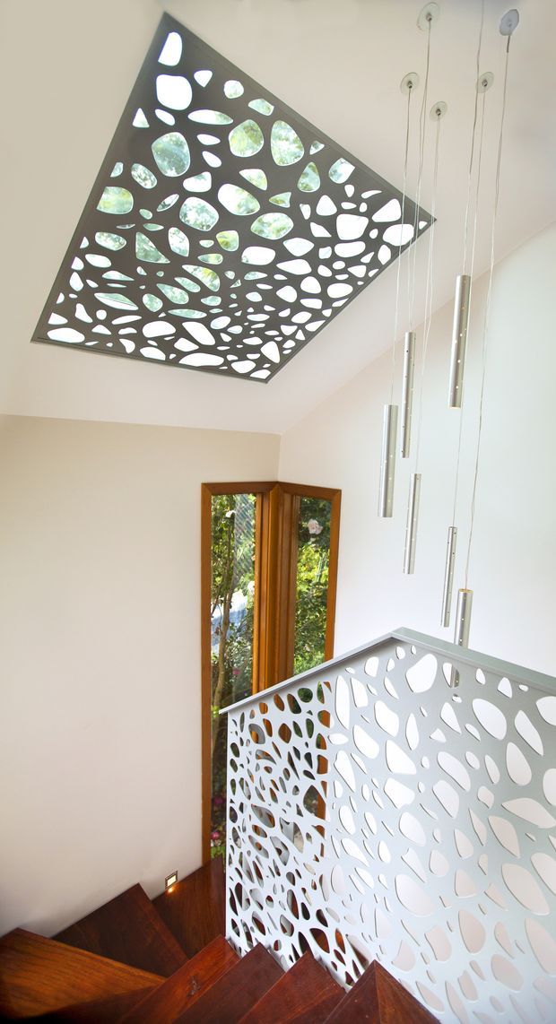 A bright contemporary stairwell viewed from above, featuring a dark timber staircase, a white perforated organic-pattern balustrade, a matching perforated ceiling panel revealing greenery beyond, suspended slim pendant lights, and timber-framed windows overlooking vegetation. The space has high ceilings and strong natural light.