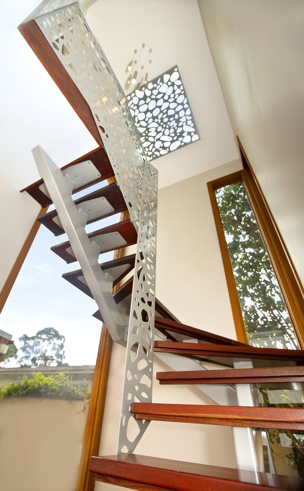 Interior view looking upward through a modern stairway with open risers, dark timber treads, slim white structural supports, and a perforated decorative metal balustrade. Tall timber-framed glazing brings in daylight and views of greenery, while a matching perforated feature panel is visible above.