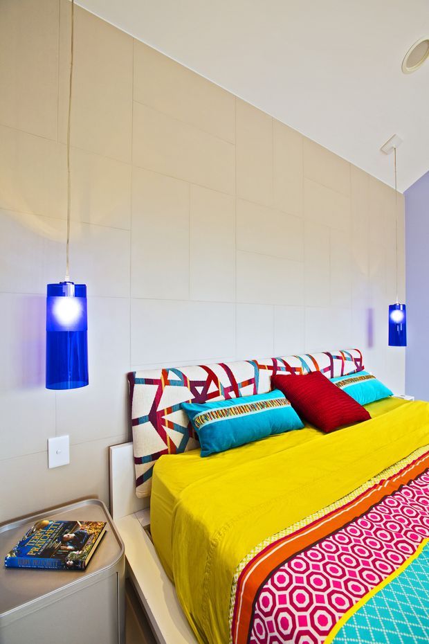 A bright, compact bedroom with a double bed dressed in vivid yellow, turquoise, red, and patterned textiles. The bed has geometric upholstered or fabric headboard panels, colourful cushions, a simple white bedside table, and striking cobalt-blue cylindrical pendant lights suspended on either side. The walls are clad in large pale beige panels, with a white ceiling and recessed spotlight. The overall look is playful, bold, and contemporary.