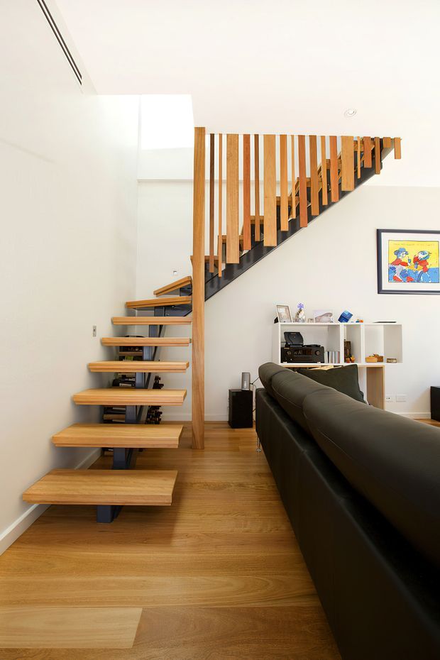 Bright contemporary interior showing a sculptural open-riser timber staircase with black steel supports and vertical timber balustrade. The staircase rises beside a living room with a dark sofa, white open shelving, timber flooring, and framed artwork. A high void above the stair adds openness and drama.