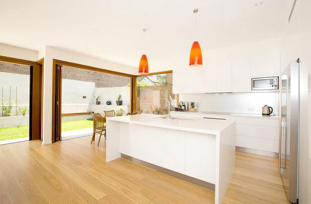 Bright contemporary kitchen with a large white island, integrated white cabinetry, stainless-steel appliances, timber-look flooring, and two vivid orange pendant lights. Wide glazed doors open to a small landscaped courtyard, bringing in abundant natural light.