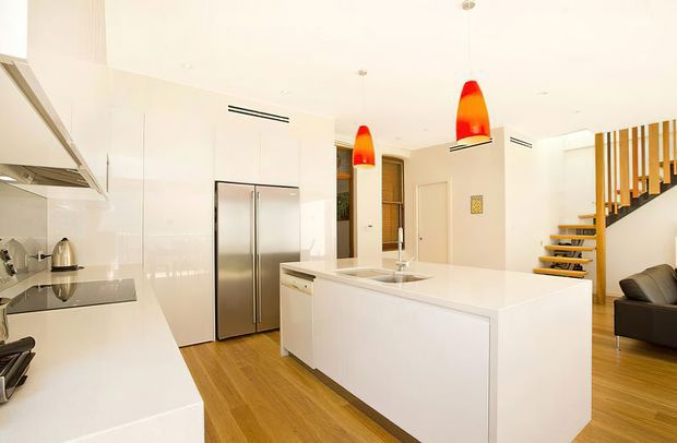 Bright contemporary open-plan kitchen with a large white island, integrated white cabinetry, stainless-steel refrigerator and appliances, timber flooring, and two striking red-orange pendant lights. An open staircase with timber treads and vertical timber balustrade is visible at the rear, along with part of a living area.
