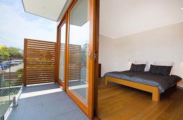 A bright modern bedroom opening onto a balcony or terrace through full-height timber-framed glazing. The room has a low-profile bed with dark bedding, warm timber flooring, white walls and ceiling, and recessed downlights. Outside, the balcony features grey tiled flooring, a clear glass balustrade, and a horizontal timber privacy screen.