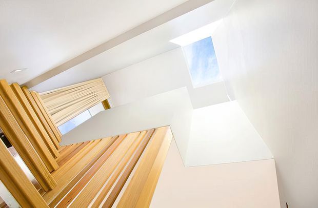 Upward-looking architectural interior view of a bright stairwell or vertical circulation space. Warm timber slatted elements and a timber-framed opening contrast with white walls and ceiling planes, while a skylight brings in strong natural light. The angled composition emphasizes the vertical void and contemporary geometric detailing.