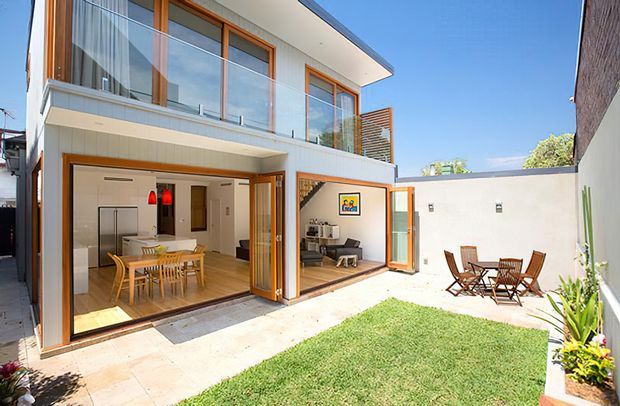 Bright contemporary two-storey residential home viewed from a private courtyard. The facade combines white rendered walls, warm timber-framed floor-to-ceiling glazing, a projecting upper level, and a timber privacy screen. Large glazed openings connect the courtyard to an open-plan dining and kitchen area and a living room, with a small lawn, paved terrace, and outdoor dining furniture.
