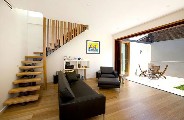 Bright, open-plan contemporary living area with a floating timber staircase and vertical timber balustrade, black leather seating, pale timber flooring, and a large opening to a sunlit courtyard or outdoor terrace. White walls and recessed ceiling lighting create a clean, spacious interior.
