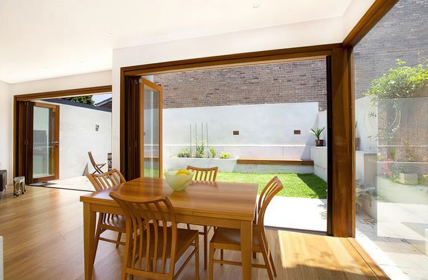 Bright dining area with a solid timber dining table and matching wooden chairs, opening through large timber-framed glazed doors to a landscaped internal courtyard. The space has warm timber flooring and joinery, white walls, and abundant natural light. A brick feature wall, raised planter beds, lawn, and outdoor seating are visible beyond the open doors.