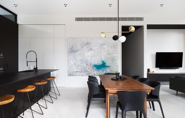 Open-plan contemporary kitchen and dining area with a dark island and bar stools, white integrated cabinetry, a timber dining table with black chairs, and a connected living area. A large abstract artwork and sculptural pendant light add visual interest, while recessed ceiling lights and linear air-conditioning vents contribute to the clean architectural fit-out.