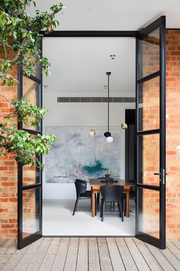 View through open black-framed glazed doors from a timber deck into a refined dining room. Exposed red brick walls, a white ceiling, pale smooth flooring, black dining chairs, a timber dining table, sculptural pendant lighting, a large abstract artwork, and indoor greenery create a contemporary industrial-leaning interior with strong indoor-outdoor connection.
