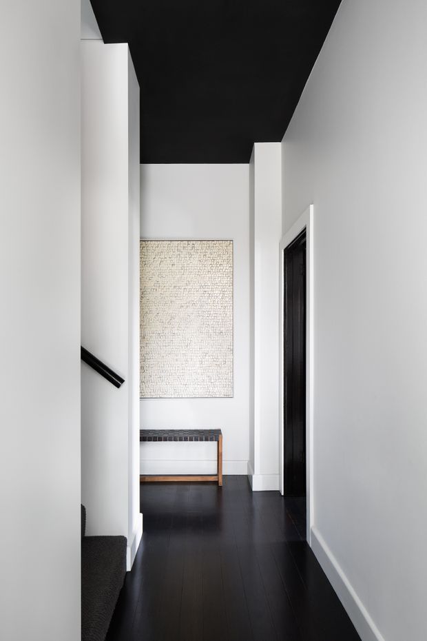 A narrow, minimalist hallway with white walls, a dramatic matte-black ceiling, dark timber flooring, and black door and stair details. A small wooden bench and a pale textured wall artwork provide restrained contrast at the end of the corridor.