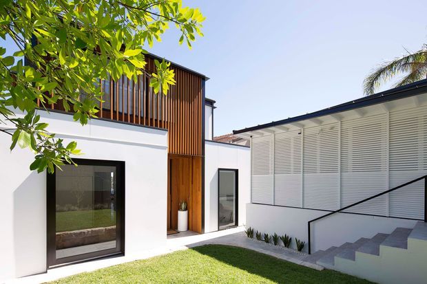 A bright contemporary residential courtyard exterior with white rendered walls, dark-framed glazing, a warm timber vertical-screen upper facade, and a low-slope dark roof. A white louvred facade and stepped stairway frame a small lawn courtyard, with tropical planting around the edges.