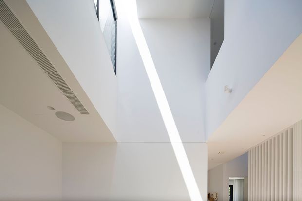 Minimalist interior view of a bright, high-ceilinged circulation space or stairwell, dominated by crisp white walls and ceiling planes. A narrow linear skylight or roof glazing casts a strong diagonal shaft of daylight across the central wall, while a vertical slatted feature wall and partial doorway are visible on the right. Built-in linear air-conditioning grilles and recessed ceiling fixtures reinforce the clean contemporary detailing.