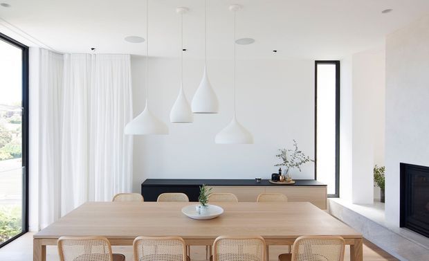 Bright, minimalist dining room with a long light-wood dining table and woven-back chairs. The room features floor-to-ceiling glazing with sheer curtains, a white ceiling and walls, multiple sculptural white pendant lights, a low dark-wood sideboard, and a contemporary fireplace set into a pale stone surround.