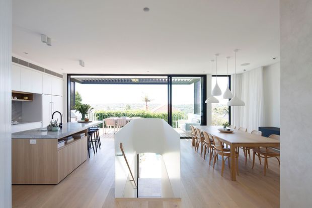 Bright open-plan residential interior combining a contemporary kitchen, dining area and living space, with a central stair opening and expansive black-framed glazing leading to an outdoor terrace. The kitchen has white upper cabinetry, warm timber lower cabinetry and a dark stone benchtop. A long timber dining table with matching chairs sits beneath multiple white pendant lights. Light timber flooring, white walls and sheer curtains create an airy, refined atmosphere, while a distant landscaped view is visible beyond the glazing.