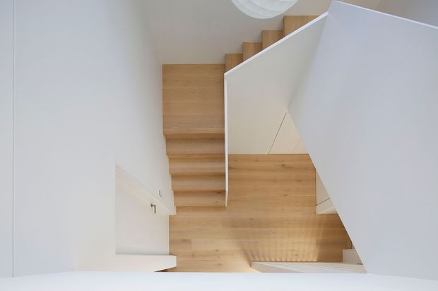 Top-down view of a minimalist residential stairway with light timber treads and matching timber flooring, enclosed by crisp white walls and a sculptural white balustrade. The stair turns around a central landing, with a simple round ceiling light partially visible above.