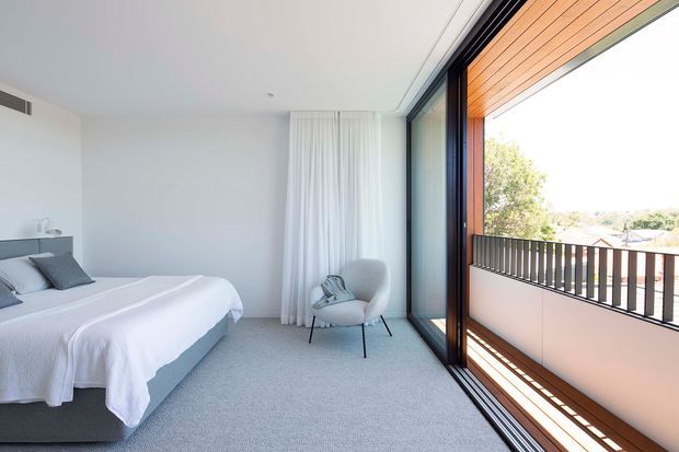 Bright, minimalist bedroom with a low upholstered bed dressed in white linens, a pale grey lounge chair, full-height white curtains, and soft grey carpeting. Large black-framed sliding glass doors open to a balcony with timber soffit lining and a dark vertical-slat balustrade. The room has a clean, airy contemporary feel with views to leafy surroundings.