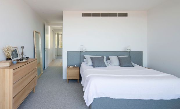 Bright contemporary bedroom with a neatly made double bed, upholstered grey headboard, pale grey carpet, white walls, and light timber bedside and dresser furniture. A hallway and doorway are visible beyond the room, with a full-length mirror and simple decorative accessories. The room has a calm, clean, softly neutral aesthetic.