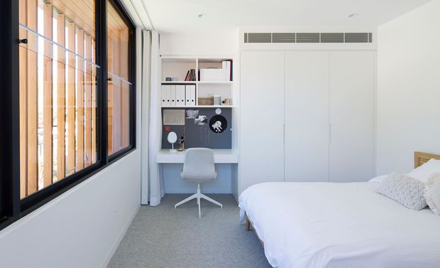 Bright, compact bedroom with a built-in study nook and full-height white storage cabinetry. The room has pale grey carpet, white walls and ceiling, a simple light timber bed frame, and a large black-framed window with vertical timber screening and curtains. The study area includes fixed white shelving and cabinetry, a grey desk surface, and a white swivel chair. The overall design is clean, understated, and contemporary.