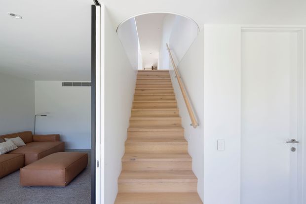 Bright contemporary interior view of a light timber staircase ascending through a curved white opening. A simple timber handrail runs along the right wall, with a white door beside the stair and a partially visible living room with a brown sectional sofa to the left. The space has clean white walls, pale timber stair treads, and a minimal, airy appearance.